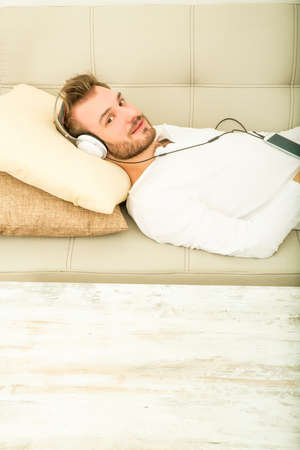 A young man lying on the sofa listening to music with some headphones and a smartphone.の写真素材