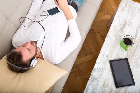 A young man lying on the sofa listening to music with some headphones and a smartphone.の写真素材