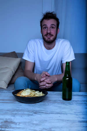 A young man watching with passion TV at nighttime with potato chips and beer at home in the living room.の写真素材