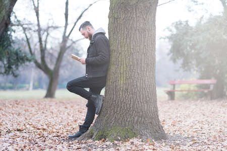 A handsome young hipster man leaning against a tree and reading a book in the park.