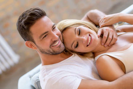 A beautiful young woman lying on the sofa with boyfriend hugging her from behind.