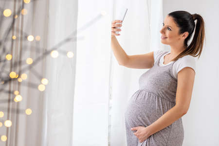 A young pregnant woman standing next to the window in an elegant grey dress and taking a selfie with...