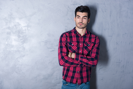 A serious handsome young man in a red checkered shirt standing in front of a grey wall in a studio.の写真素材