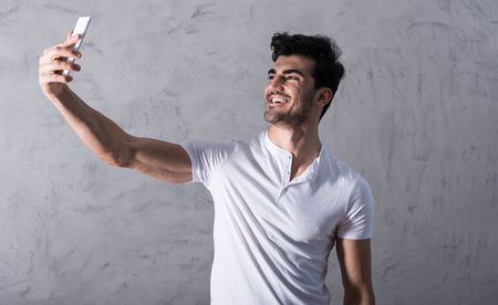 A happy handsome young man in a white tshirt taking a selfie with his smartphone in front of a grey wall in a studio.の写真素材