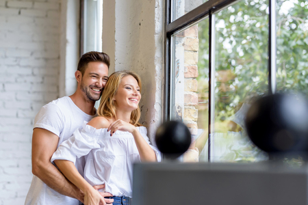 A beautiful young woman standing next to the window with boyfriend hugging her from behind.の写真素材