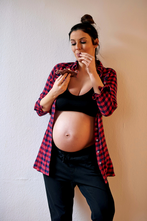 A young happy pregnant woman in a red shirt and a black underwear eating a cupcake in front of a grey background in a studio.の写真素材