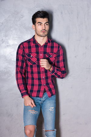 A serious handsome young man in a red checkered shirt standing in front of a grey wall in a studio.の写真素材