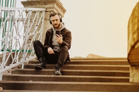 A handsome young hipster man sitting on the stairs and taking a selfie with his smartphone.の写真素材
