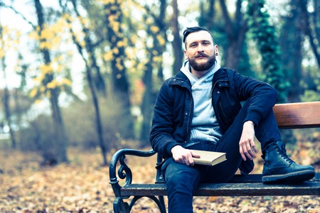 A retro style photo of a young hipster man sitting on a bench with a book and daydreaming.