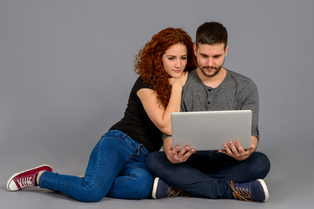 A beautiful young couple sitting on the ground and looking at a laptop and smilingの写真素材