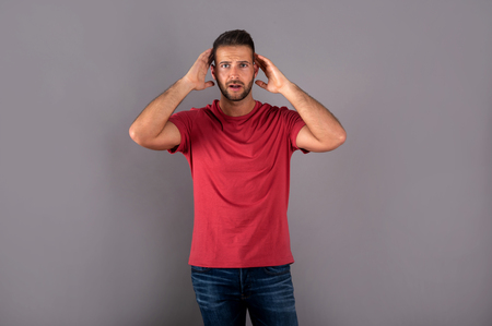 An upset handsome young man in a red tshirt standing in front of a grey background in the studio.の写真素材