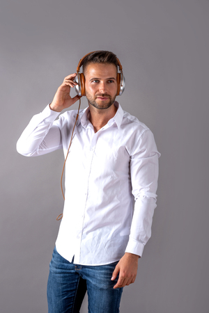 A serious handsome young man in a white shirt listening to music on his headphones and standing in front of a grey background in the studio.の写真素材