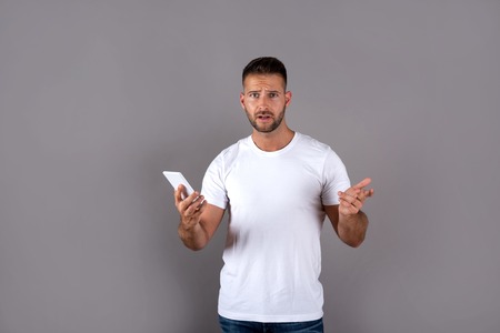 A worried handsome young man in a white tshirt using his smartphone and standing in front of a grey...