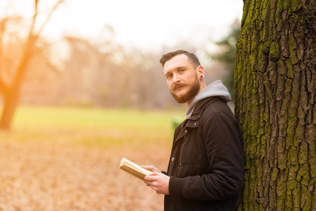 A smiling handsome young hipster man leaning against a tree and reading a book in the park.の写真素材