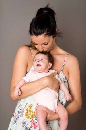 A beautiful young mother standing and holding her baby girl in her arms in front of a grey background in a studio.の写真素材