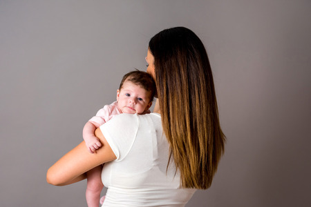 A beautiful young mother standing and holding her baby girl in her arms in front of a grey background in a studio.の写真素材