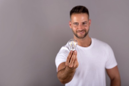 A smiling young man in a white tshirt holding a lightbulb and standing in front of a grey background in the studio.の写真素材