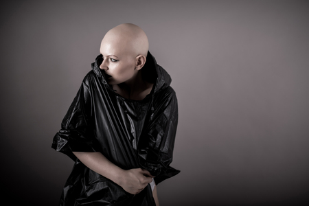 Emotive photo of a beautiful bald woman while standing in the studio and wearing a black raincoat.の写真素材