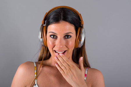 A beautiful surprised young woman listening to music with a pair of headphones in front of a grey background in a studio.の写真素材