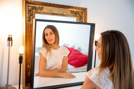 Portrait of a beautiful mature woman in a white dress while sitting in front of a mirror in a modern apartments living room.の写真素材