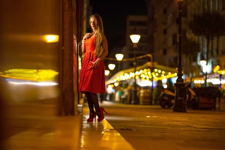 A beautiful mature woman in a red dress checking the storefront at night with the city lights in the background.の写真素材