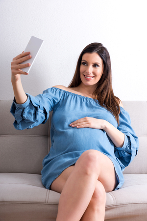 A beautiful young pregnant woman sitting on a sofa in an elegant blue dress and taking a selfie with her smartphone.の写真素材