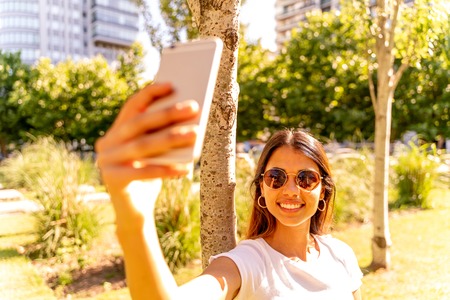 A beautiful young woman in a white shirt and sunglasses standing next to a tree and taking a selfie.の写真素材