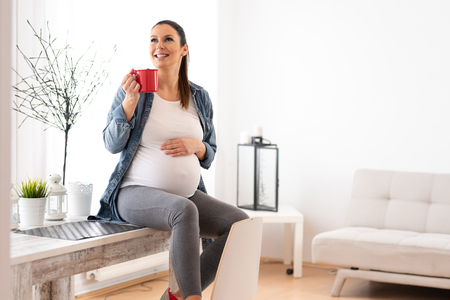 A beautiful smiling pregnant woman sitting on a kitchen table in a denim jacket and drinking a coffee from a red mug.の写真素材