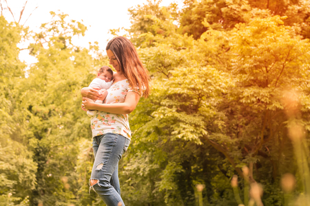 A beautiful young mother standing in the park and holding her baby girl in her arms on a sunny day.の写真素材