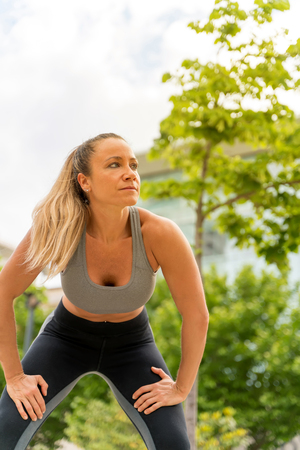 An athletic woman in a grey top standing with her hands on her knees in the park with modern buildings in the background.の写真素材