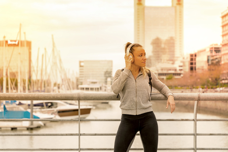 An athletic woman in a grey sweater standing next to the bridge railing and listening to music on her headphones.の写真素材