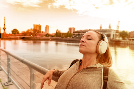 An athletic woman in a grey sweater standing next to the bridge railing and listening to music on her headphones.の写真素材
