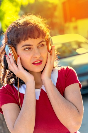 A beautiful woman in a red dress listening to music on her headphones in an urban environment.