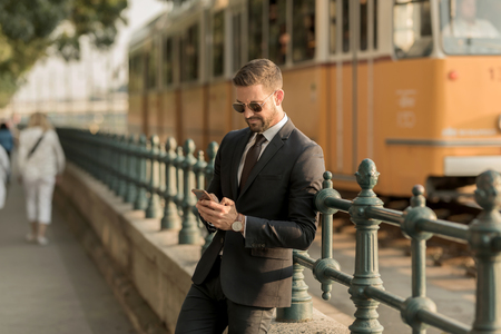 A handsome young businessman standing next to a tramline and using his smartphone on a sunny day.の写真素材