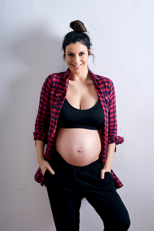 A young happy pregnant woman in a red shirt and a black underwear standing in front of a grey background in a studio.の写真素材