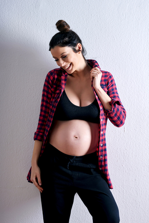 A young happy pregnant woman in a red shirt and a black underwear standing in front of a grey background in a studio.の写真素材