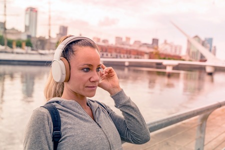 An athletic woman in a grey sweater standing next to the bridge railing and listening to music on...