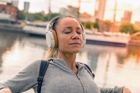 An athletic woman in a grey sweater standing next to the bridge railing and listening to music on her headphones.の写真素材