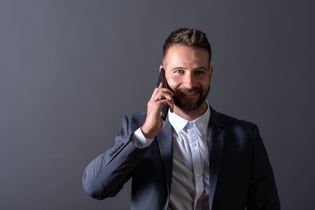 A smiling handsome young businessman in a suit talking on his phone in front of a dark grey background in a studio.の写真素材