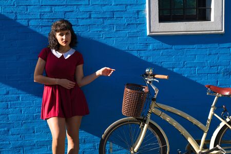 A beautiful woman in a red dress standing and posing with a bicycle in front of a brick wall on a sunny day.の写真素材