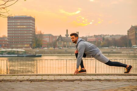 A handsome young man stretching and warming up on the riverside with the sun setting behind himの写真素材