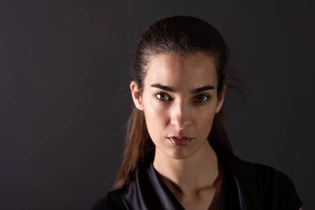 A beautiful serious young woman in an elegant black dress standing in front of a grey background in a studio.の写真素材