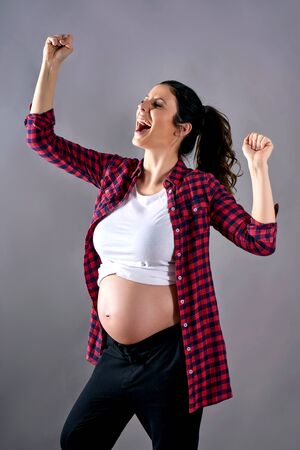 A young cheering pregnant woman in a red shirt and a white tshirt standing in front of a grey background in a studio.の写真素材