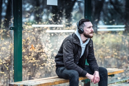 A handsome young hipster man waiting at the bus stop while listening to music on his headphones.の写真素材