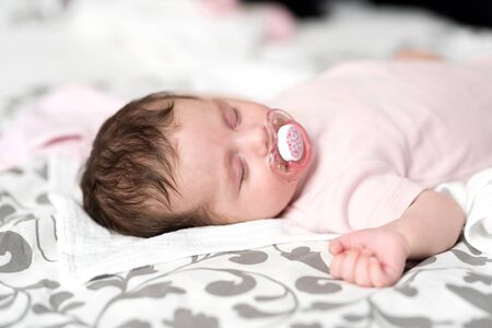 Closeup photo of a baby girl lying on a bed and sleeping with a dummy in her mouth.