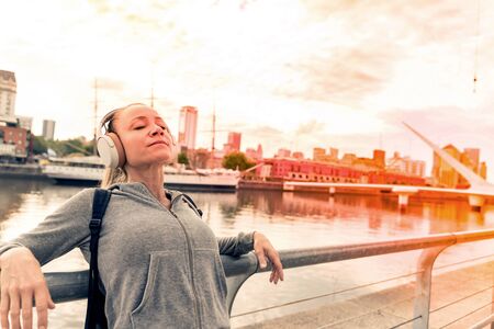 An athletic woman in a grey sweater standing next to the bridge railing and listening to music on her headphones.の写真素材