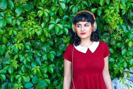 A serious woman in a red dress listening to music on her headphones in front of green leaves on a...