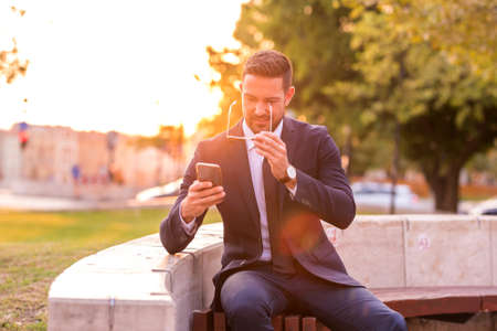 A handsome young businessman sitting on a bench and using his phone on a sunny day.