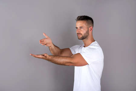 A handsome young man in a white tshirt showing something with his palms in front of a grey background in the studio.の写真素材