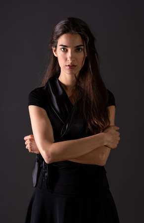 A beautiful serious young woman in an elegant black dress standing in front of a grey background in a studio.の写真素材